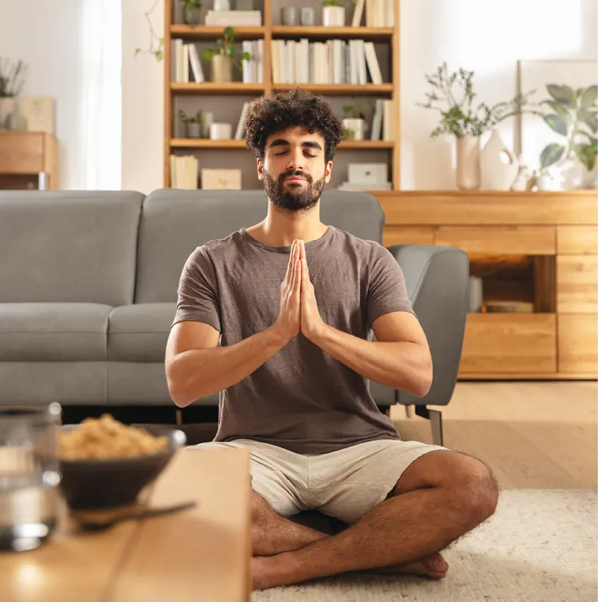 Mann sitzt meditierend im Wohnzimmer vor einem grauen Sofa und Holzmöbeln. Ruhige Wohnatmosphäre mit natürlichen Materialien, modernes Wohnen mit Komfort und stilvoller Einrichtung für mehr Entspannung im Alltag.